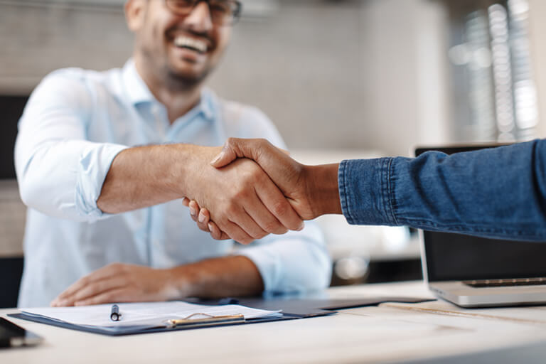 Closeup of a handshake between two people across a desk. The man on the left wears glasses and is smiling. The person on the right isn't visible except for his hand and arm. He wears a blue denim shirt.