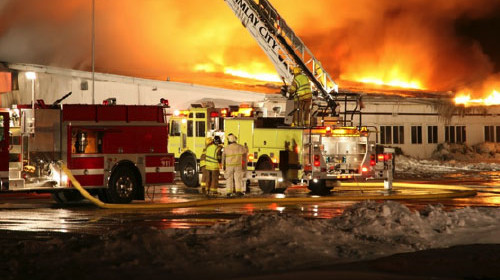 firetrucks and firefighters at scene of facility fire, flames coming out of roof