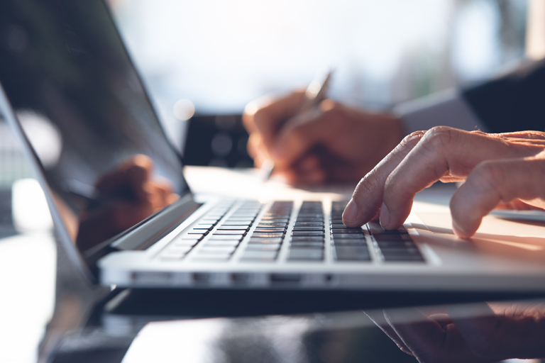 Closeup of businessman typing on a laptop keyboard with his left hand while writing on a pad with his right hand